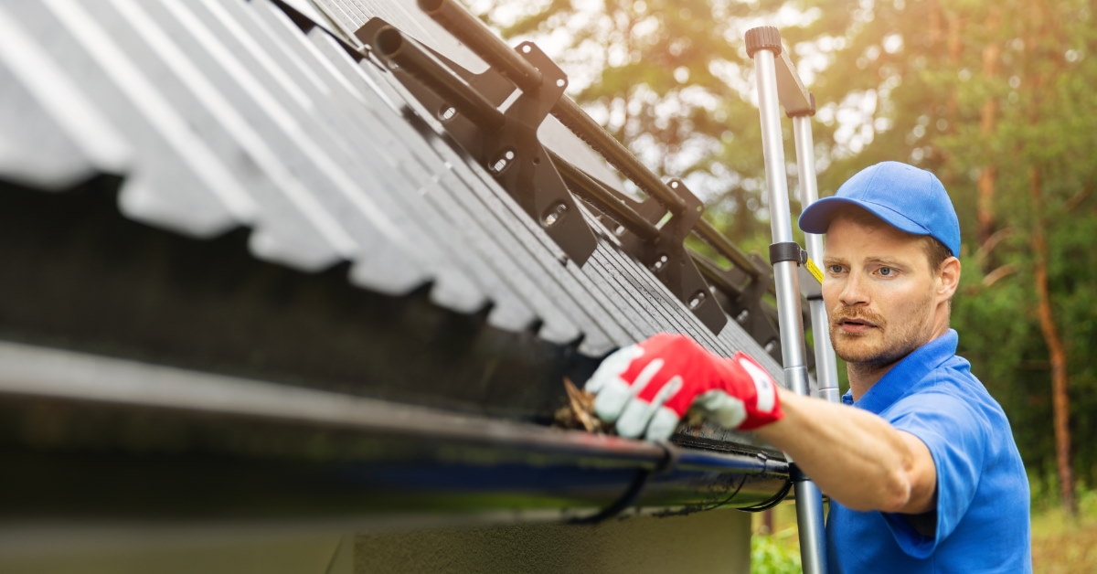 male worker wearing gloves standing on ladder clearing gutter 