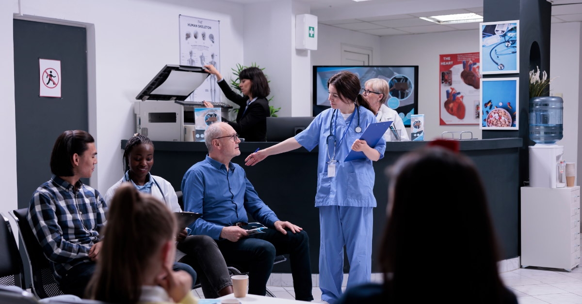female nurse holding clipboard wearing scrubs talking to senior pateint sitting in waiting area