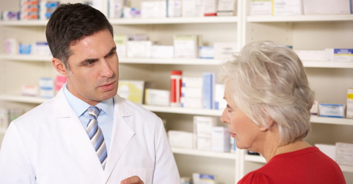 male pharmacist in pharmacy explaining medicines to senior lady in red shirt