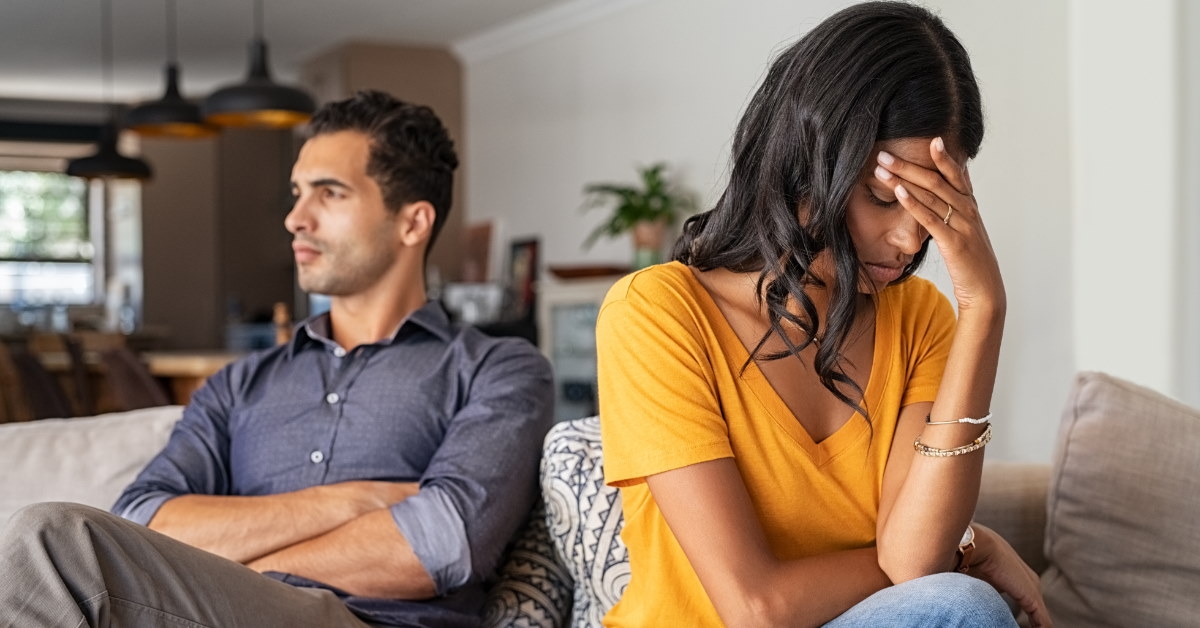 stressed wife holds her head while her husband crosses his arms after a heated argument at home