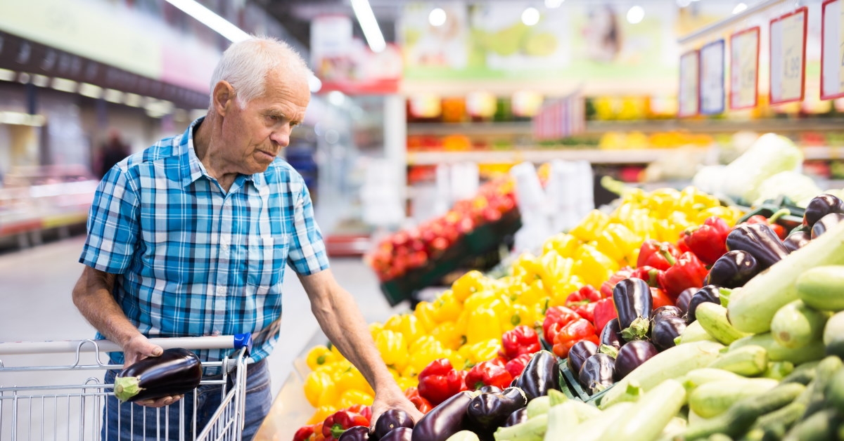 senior man standing inside grocery store buying eggplants