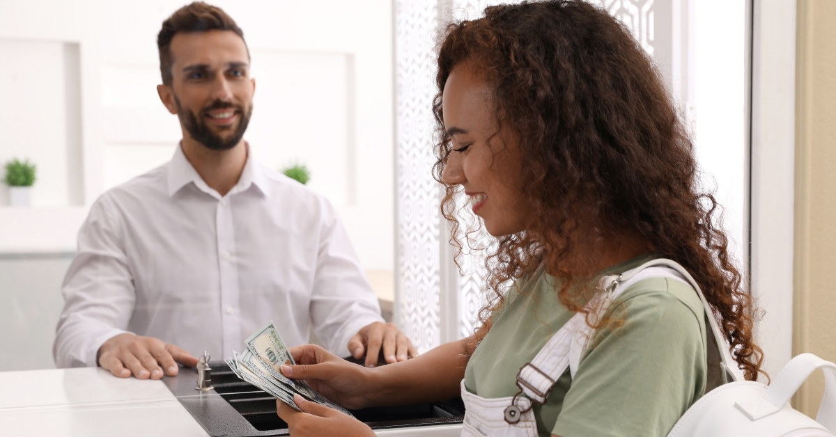 happy african american woman counting money in bank window with male cashier inside the cabin