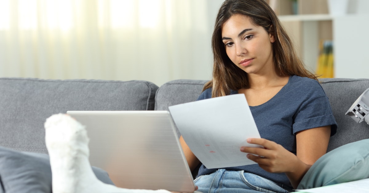 woman reviewing papers sitting on couch having plaster on feet working from home using laptop 