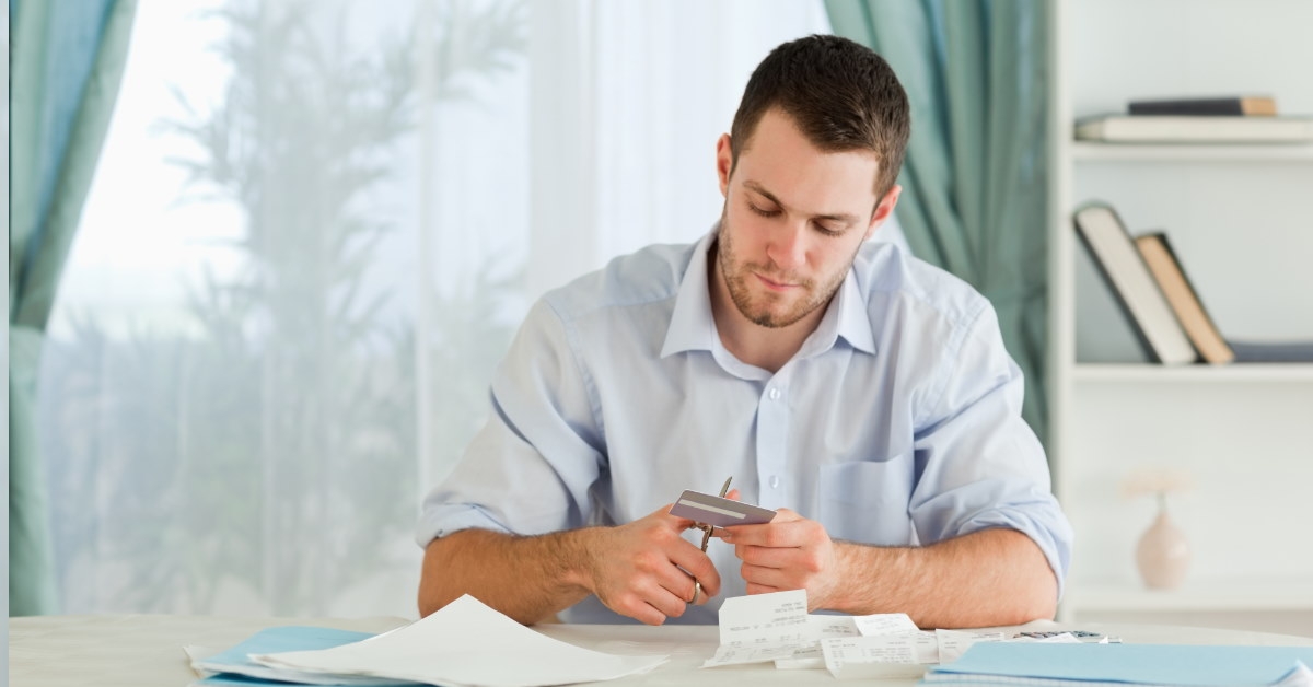 young man sitting at table with bills holding scissor to cut credit card in half
