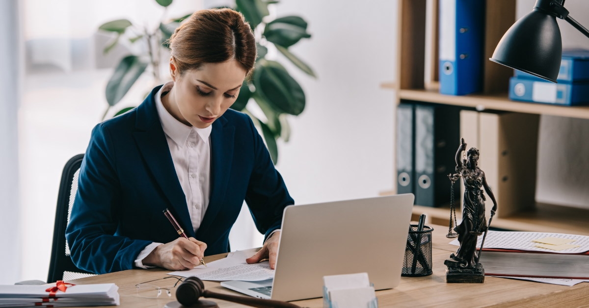 female lawyer wearing suit sitting in office writing on papers while using laptop