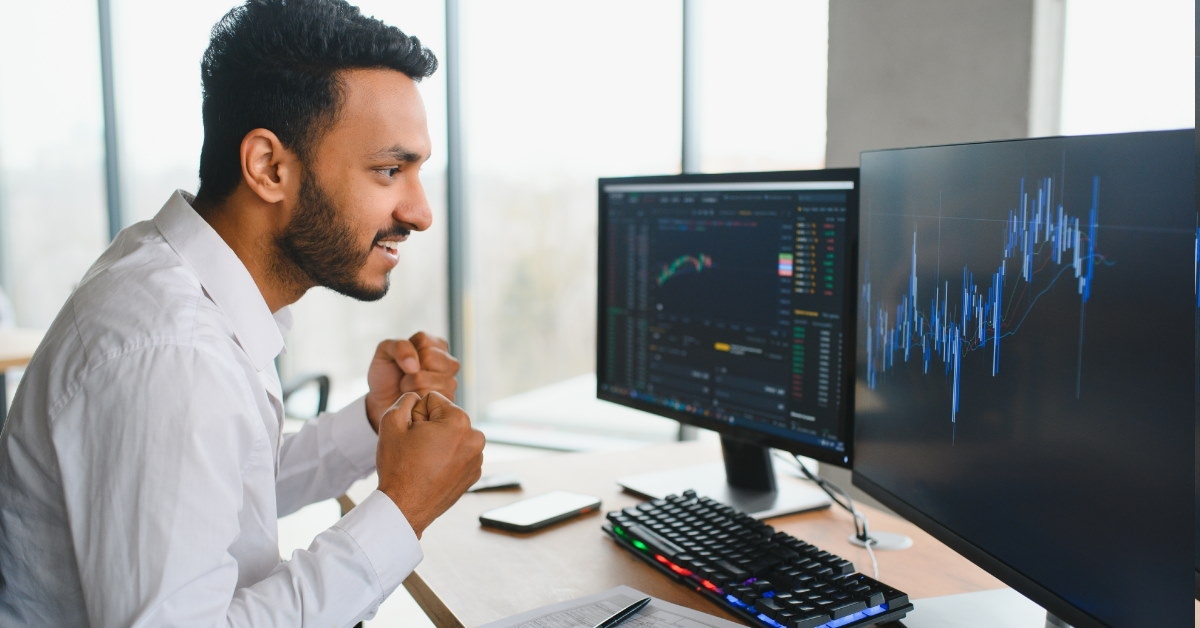 male broker sitting in front of two monitors cheering up at stock graphs