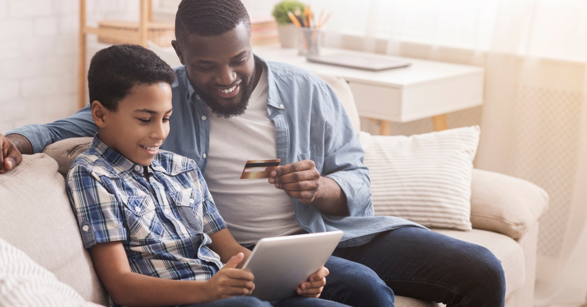 african american father and son sitting on couch holding card for online payment using tablet