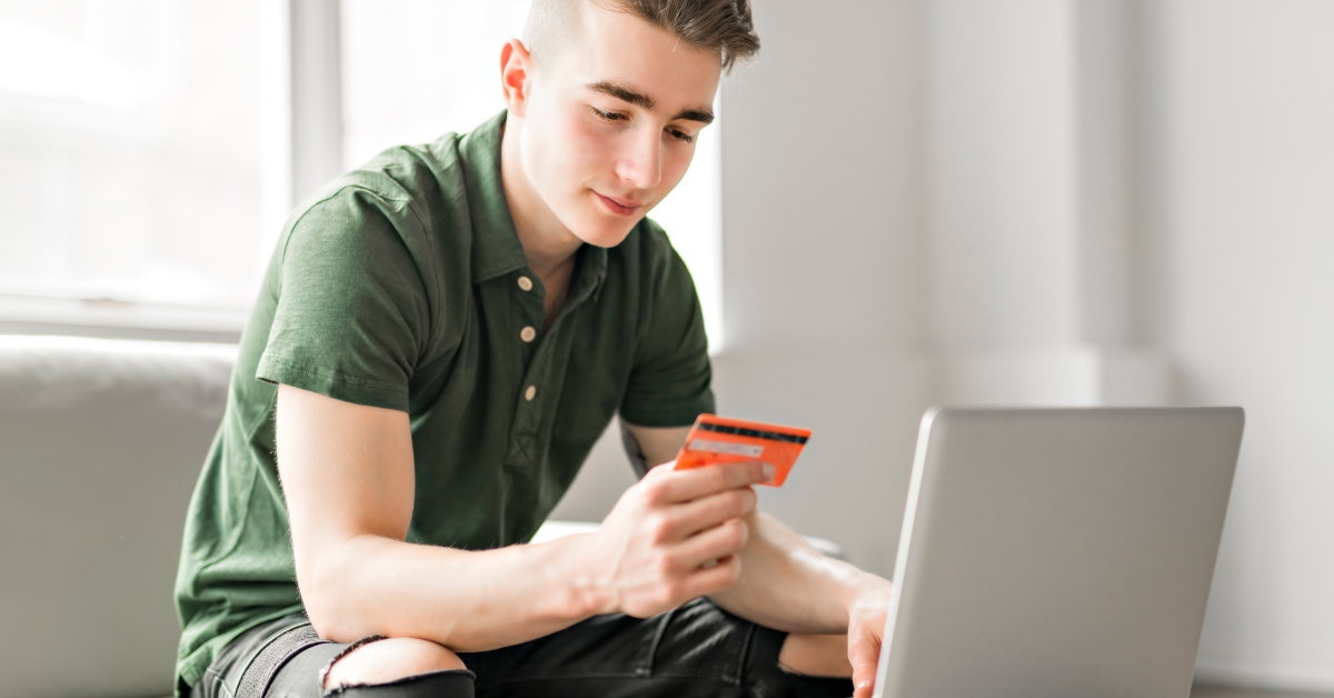 young teenage boy using laptop while holding card for making online payment at home