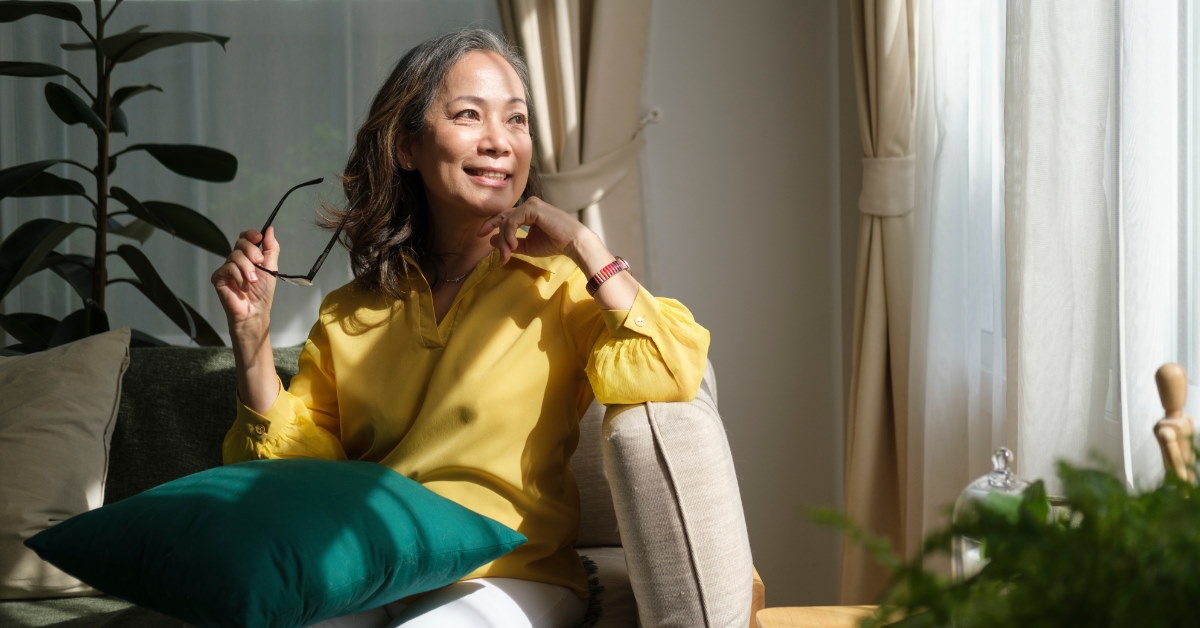 Senior woman sitting on cozy couch looking away while removing glasses at home
