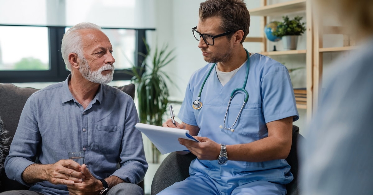 male physician wearing blue scrubs and stethoscope writing while talking to senior patient