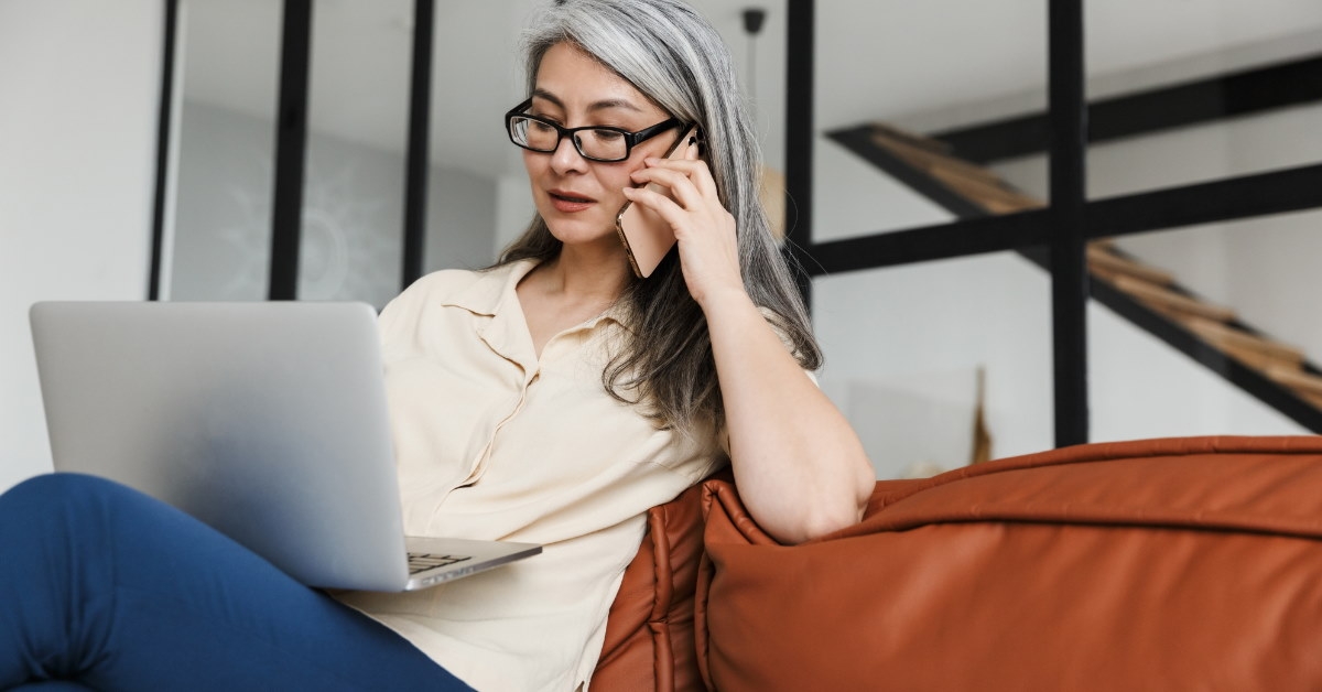 senior woman sitting on couch at home using laptop while talking on phone