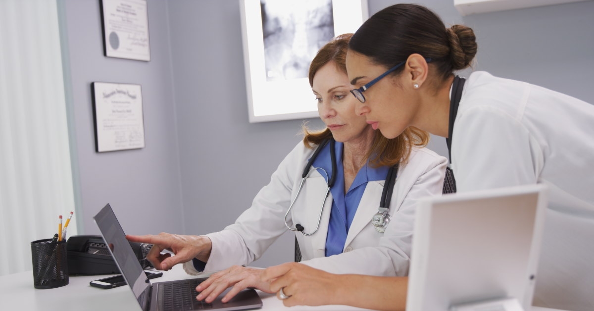 two female physician discussing case at laptop while wearing white overalls at clinic
