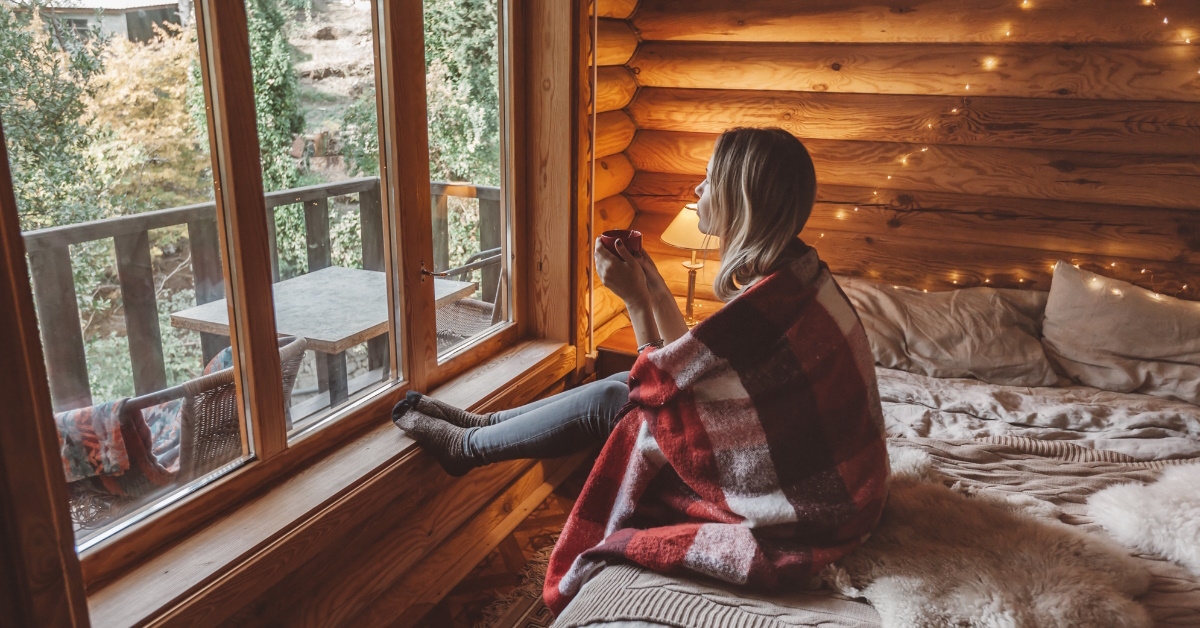 woman having cozy time while looking out of window in wooden log cabin