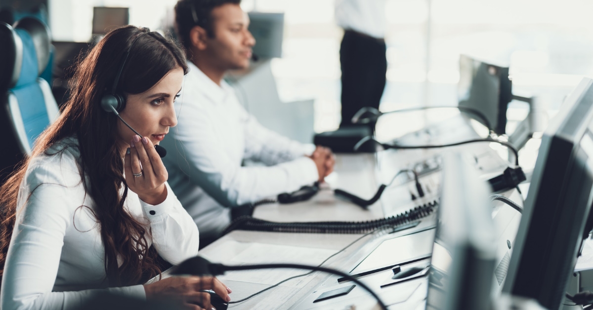 beautiful woman sitting at call center wearing headset talking to client with male colleague in background