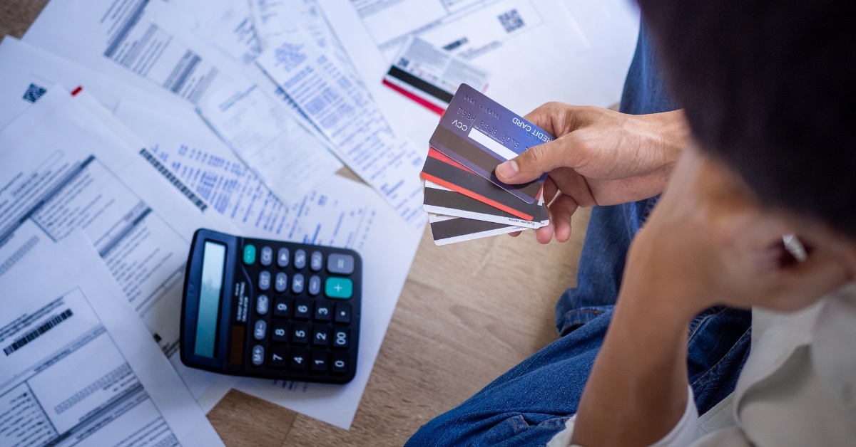 man sitting on floor with bills holding credit cards in hand thinking which one to use for payment