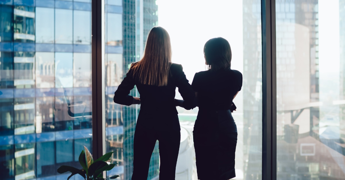 female colleagues in formal wear looking out at tall skyscrapers from office lobby