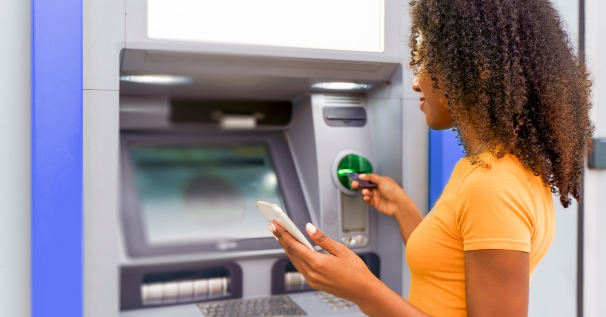 african american woman holding smartphone using card to take out money from outdoor atm