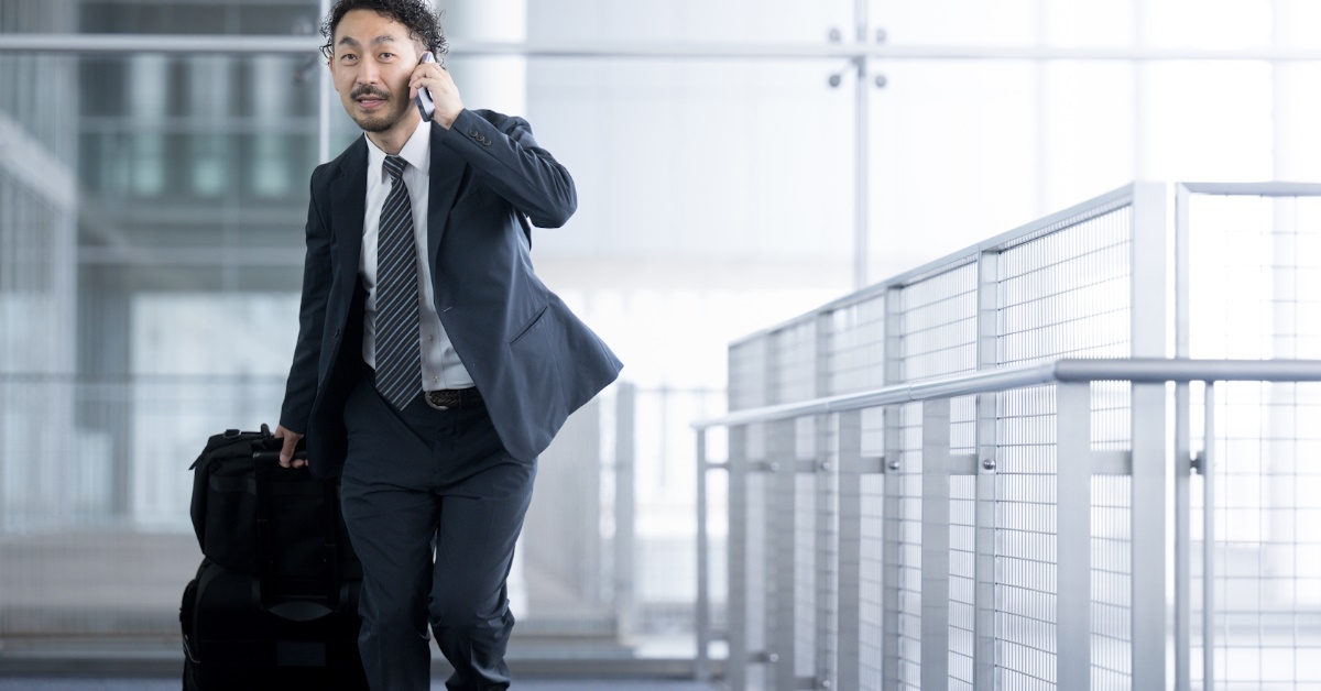 asian man walking with suitcases in airport lounge talking on smartphone