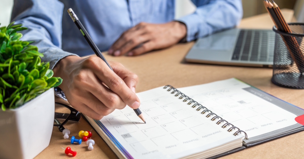 man sitting at table using pencil to mark calendar while using laptop to work from home