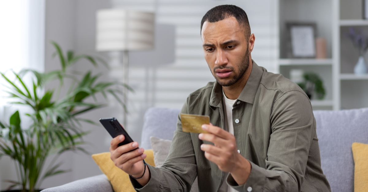Upset man sitting on sofa at home and looking at credit card