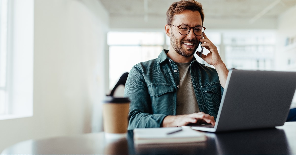male software designer working on laptop while talking to client on smartphone