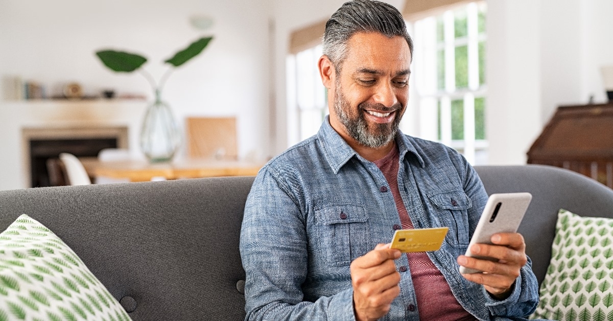 Man sitting on couch at home using card for online shopping using smartphone