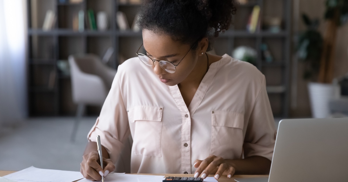 african american woman sitting at table using calculator to calculate expenses in front of laptop