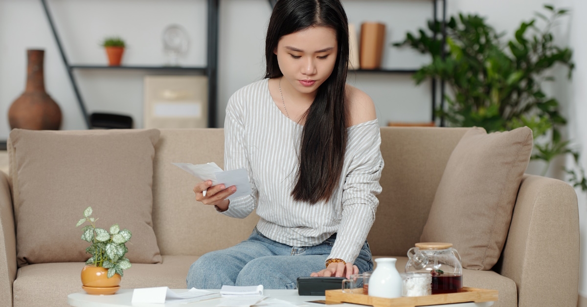 young asian woman sitting on sofa at home reviewing utility bills using calculator at table