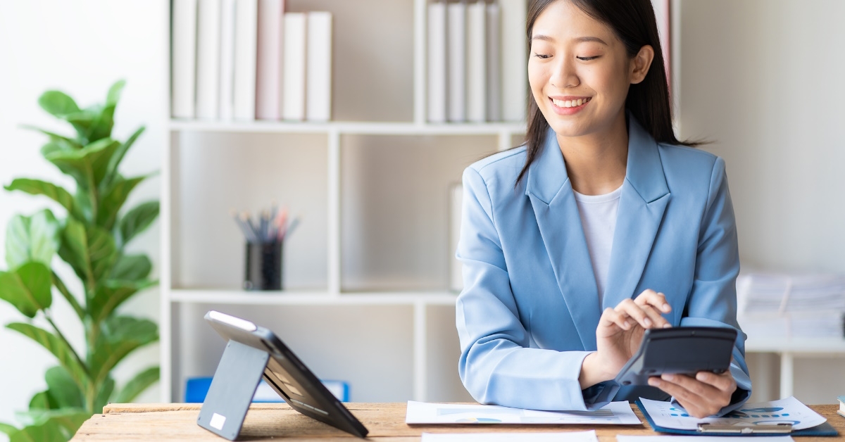 female accountant sitting at table using calculator to analyze data while looking at tablet