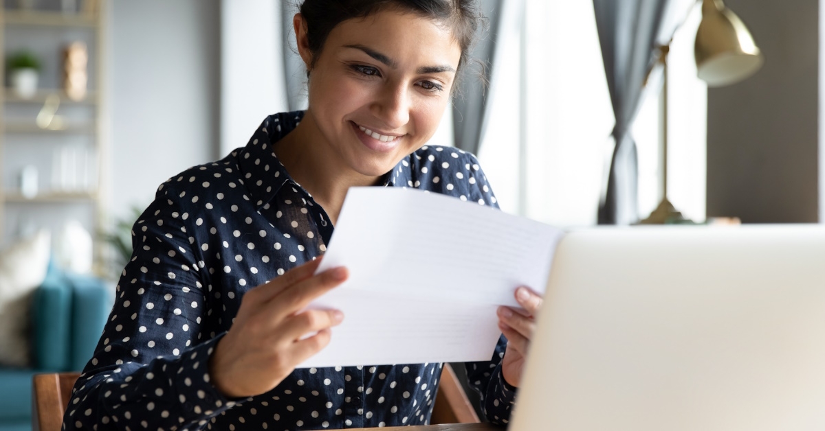 happy young woman sitting in front of laptop reading letter in hand