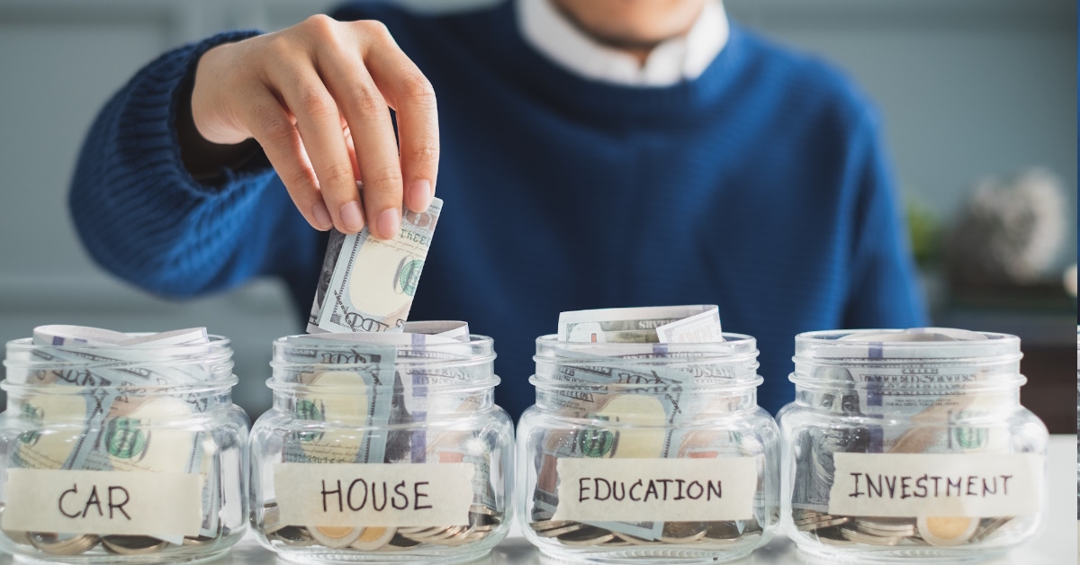 man saving money in labeled glass jars for car, house, education, and investments at a table.