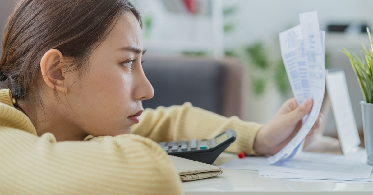 unhappy woman sitting on floor looking at receipts in hand besides calculator
