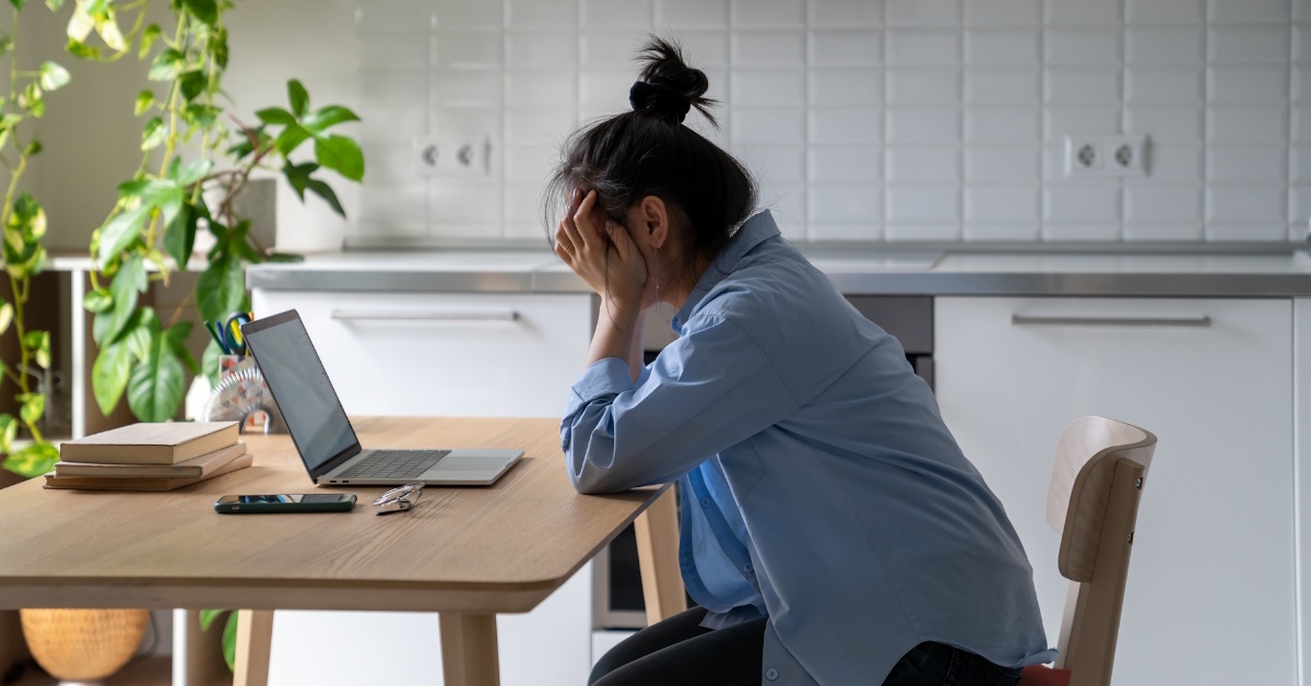 woman sitting in kitchen covering face in stress in front of laptop 