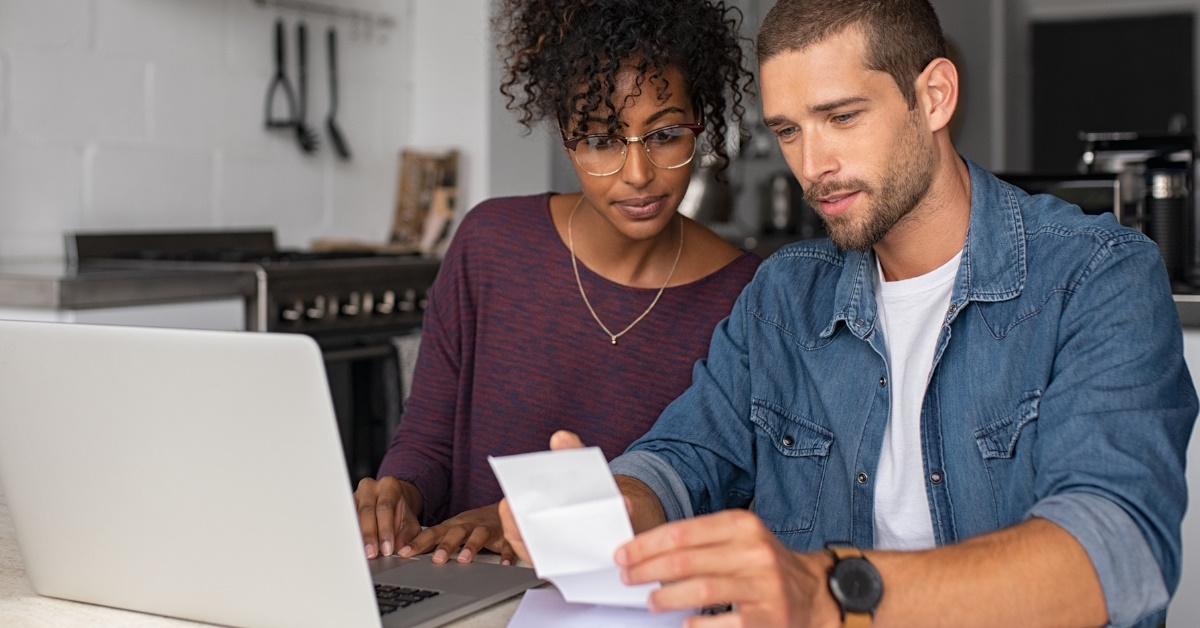 multi ethnic couple using laptop while reviewing bills in home kitchen