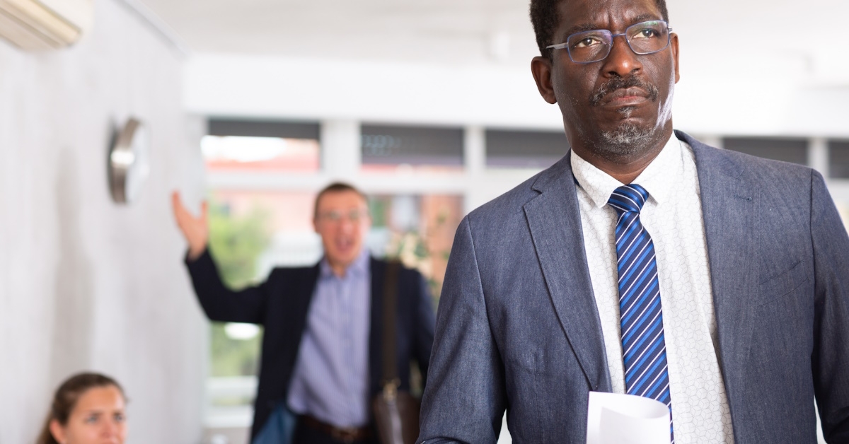 upset african american man stands in the lobby with documents while his male and female colleagues are seen in the background.