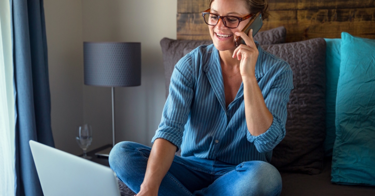 female blogger sitting on bed talking on smartphone while working on laptop