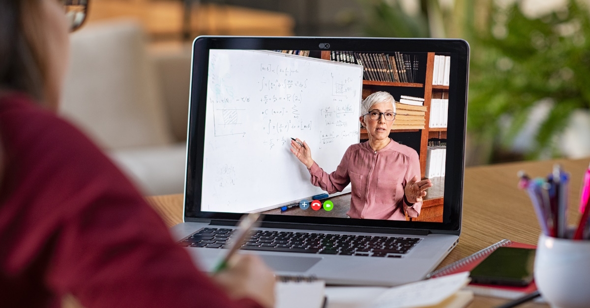 A female student is taking an online lecture on her laptop while sitting on table with her notebook and pencils.