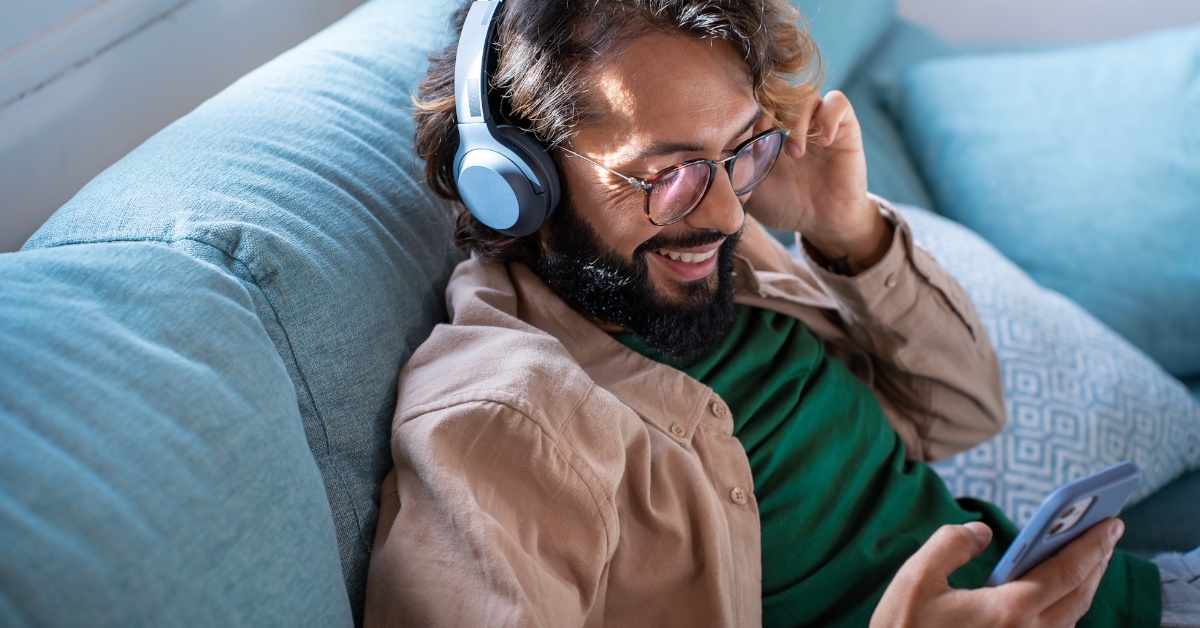 Smiling young man wearing headphones listen to mobile music playing in smartphone app while sitting on sofa ,at home.