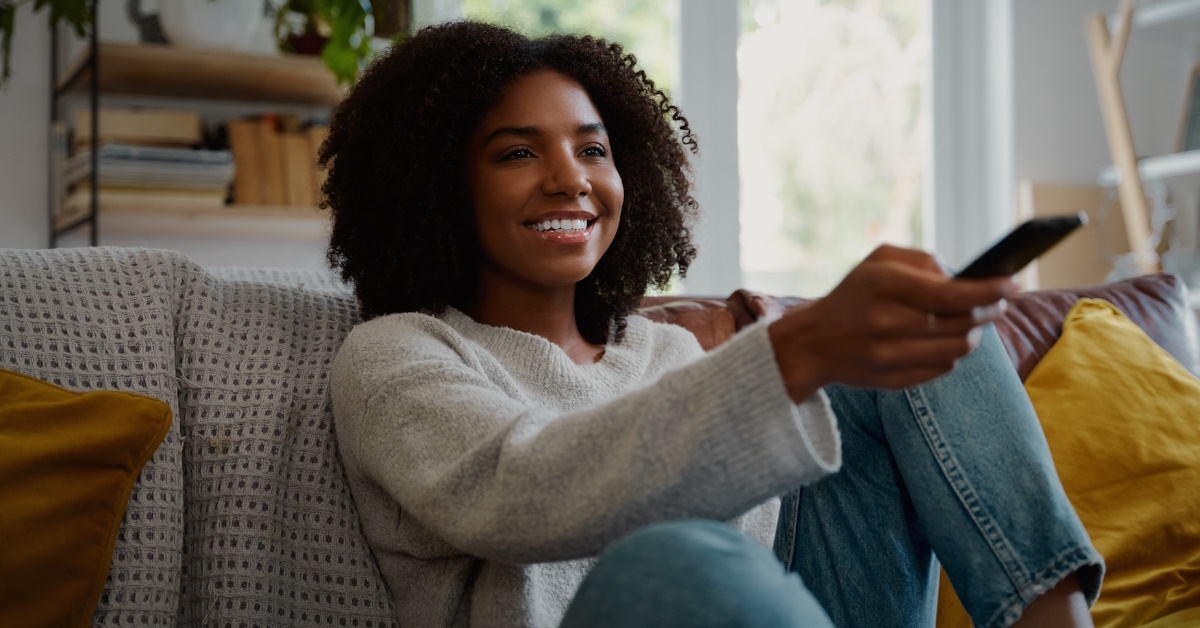 cheerful african woman sitting on couch at home watching tv and changing channels with remote control
