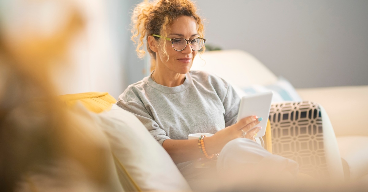 Young mature woman smiling and enjoying reading an electronic book on tablet while comfortably sitting on the sofa at home.
