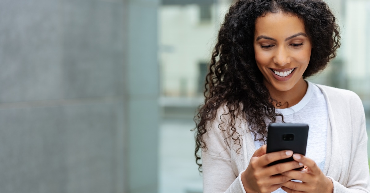 Smiling Young woman with curly hairs using a mobile while walking through town. 