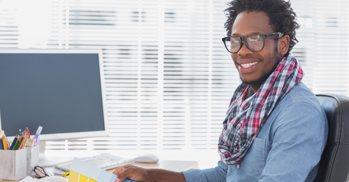 A black male interior designer is working with color charts, sitting at a table with laptops and a pencil holder