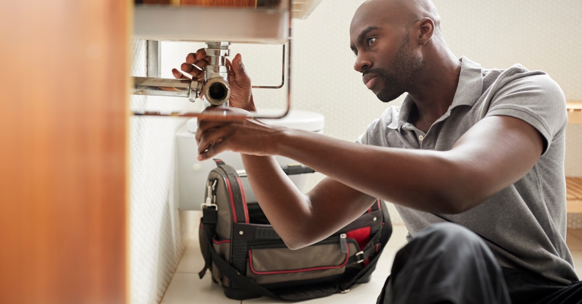  A plumber, seated on the floor, fixes a bathroom sink while viewed from the doorway.