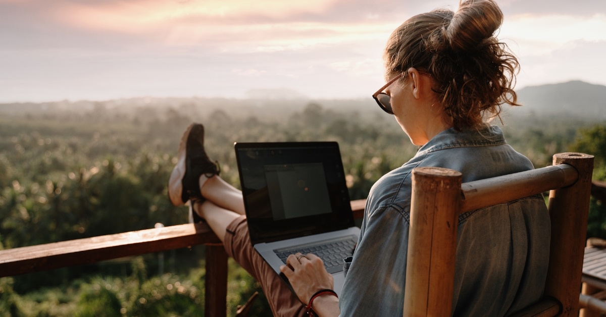 A young girl is working on her laptop while seated on a balcony in a natural environment during sunset.