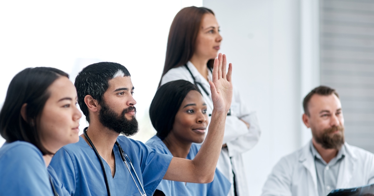A young male doctor is raising his hand during a hospital boardroom meeting and asking questions in an effort to understand the problem better.