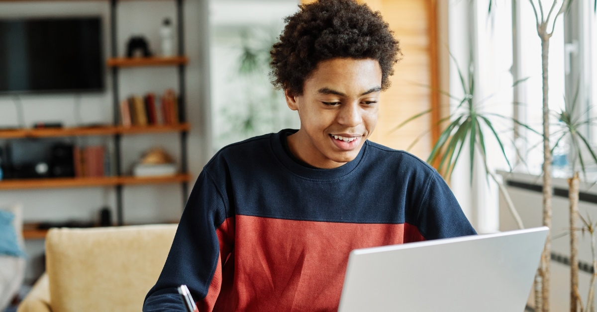 A teenage boy with curly hair is attending online classes by using his laptop while sitting in the lounge of his home.