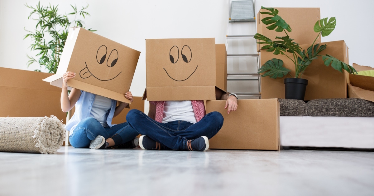 A young couple, wearing cardboard boxes with funny emojis on them, are celebrating and enjoying their move to a new home.