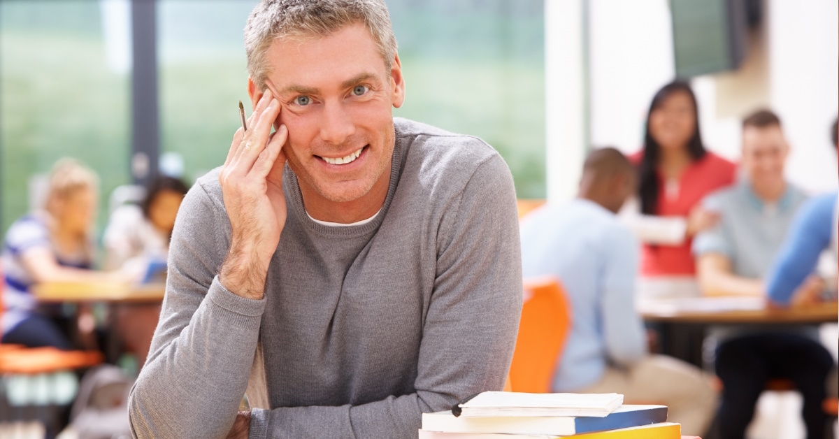 A mature male student studying with books in a library, while other students in the background engage in group study and discussion.