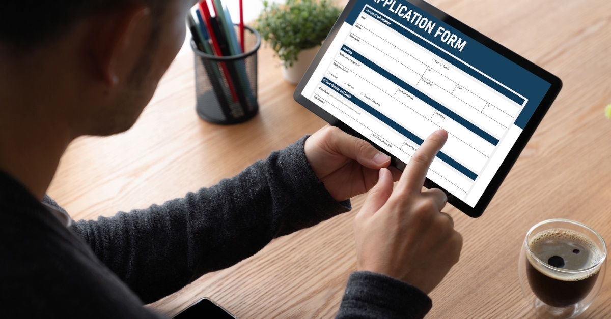 A young man is using his tablet to fill out an online job application form while sitting on table with pencil holder and a cup of tea.
