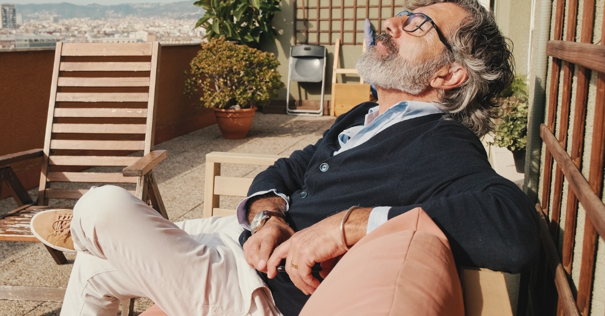 A middle-aged man, wearing glasses, sitting on a chair on the balcony and enjoying the sunny weather.